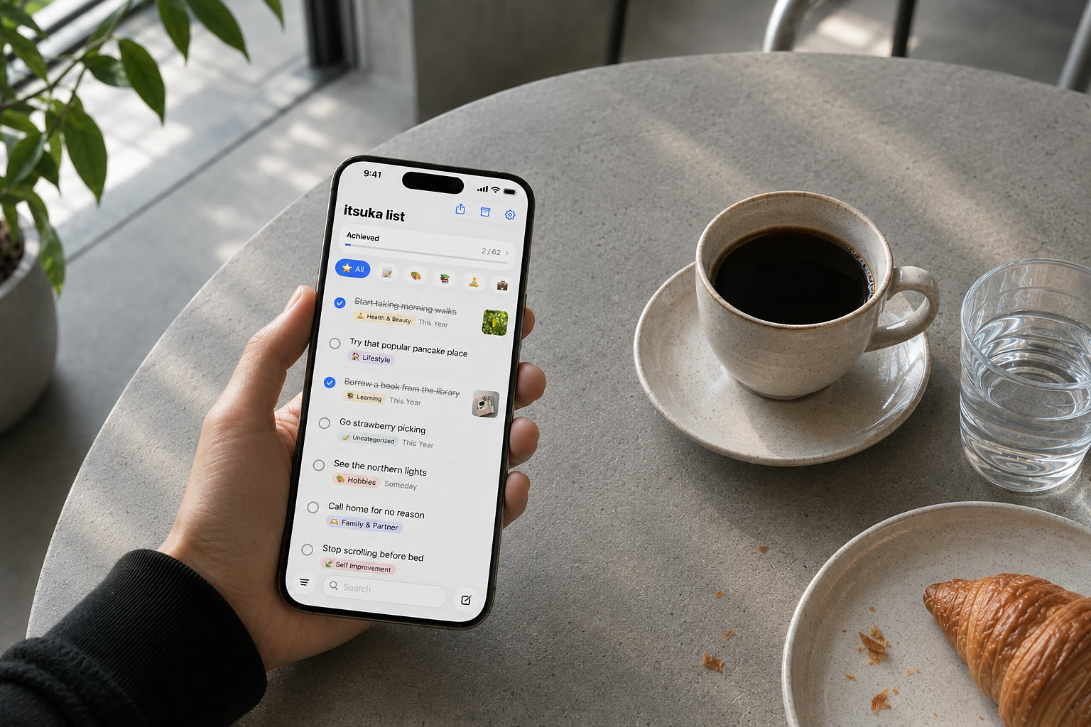 A hand holds an iPhone showing an itsuka list at a café table. A cup of coffee and a croissant are on the table.