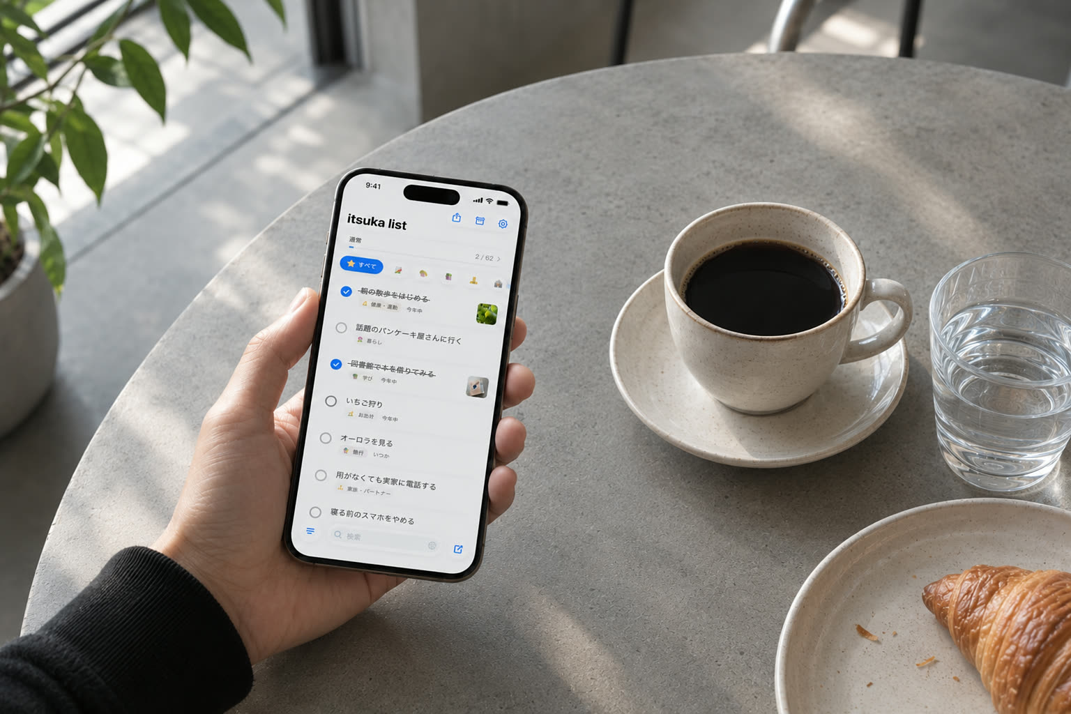 カフェの席で、itsukaのリストを表示したiPhoneを手に持っている。机にはコーヒーとクロワッサン。
